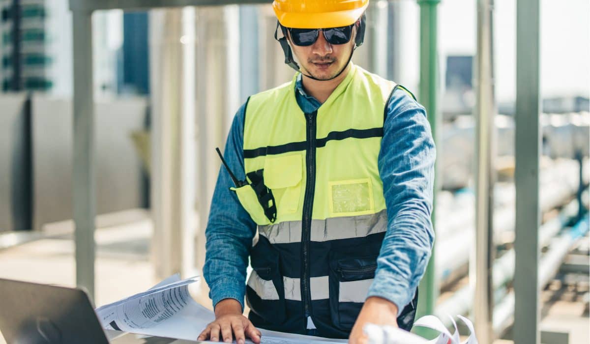 Technician in hard hat , sunglasses and high-vis jacket looking over project plans